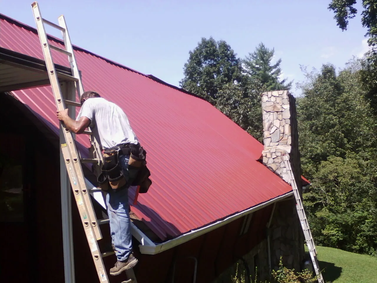 Roofer on a ladder beside a red metal roof and stone chimney — Soffit & Fascia Repair by Roofing Brothers in Langeloth